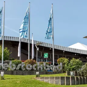 Eingangsschild „Rebstockbad“ vor Schwimmbad mit Fahnen und Grünanlage