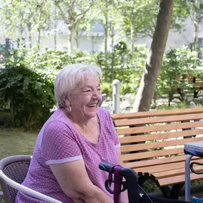 Lachende Seniorin im Garten, sitzt am Tisch mit Rollator neben Parkbank